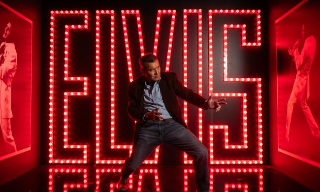 man dances like elvis in front of red neon "E-L-V-I-S" sign